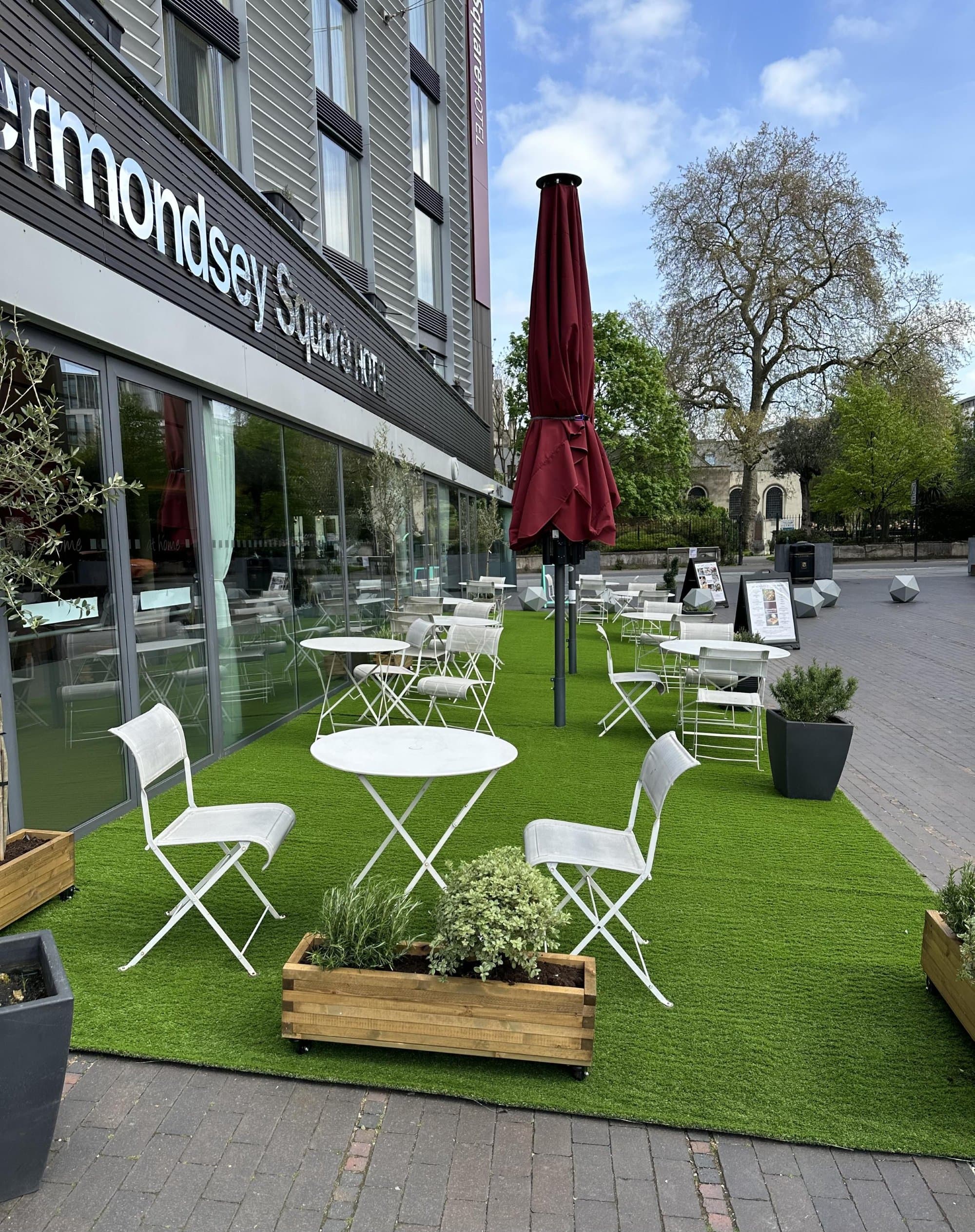 Wide finished view of the ground-floor street seating along the hotel front with red umbrellas up, café seating set out and dark planters at intervals