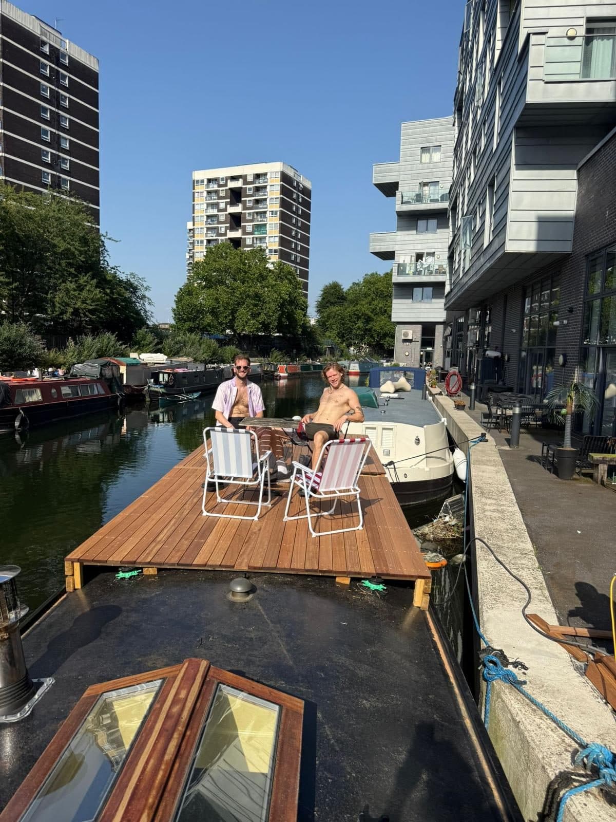 The finished hardwood roof deck on a narrowboat at Angel, two people relaxing in deck chairs at deck level with the canal, moored boats and waterside flats behind