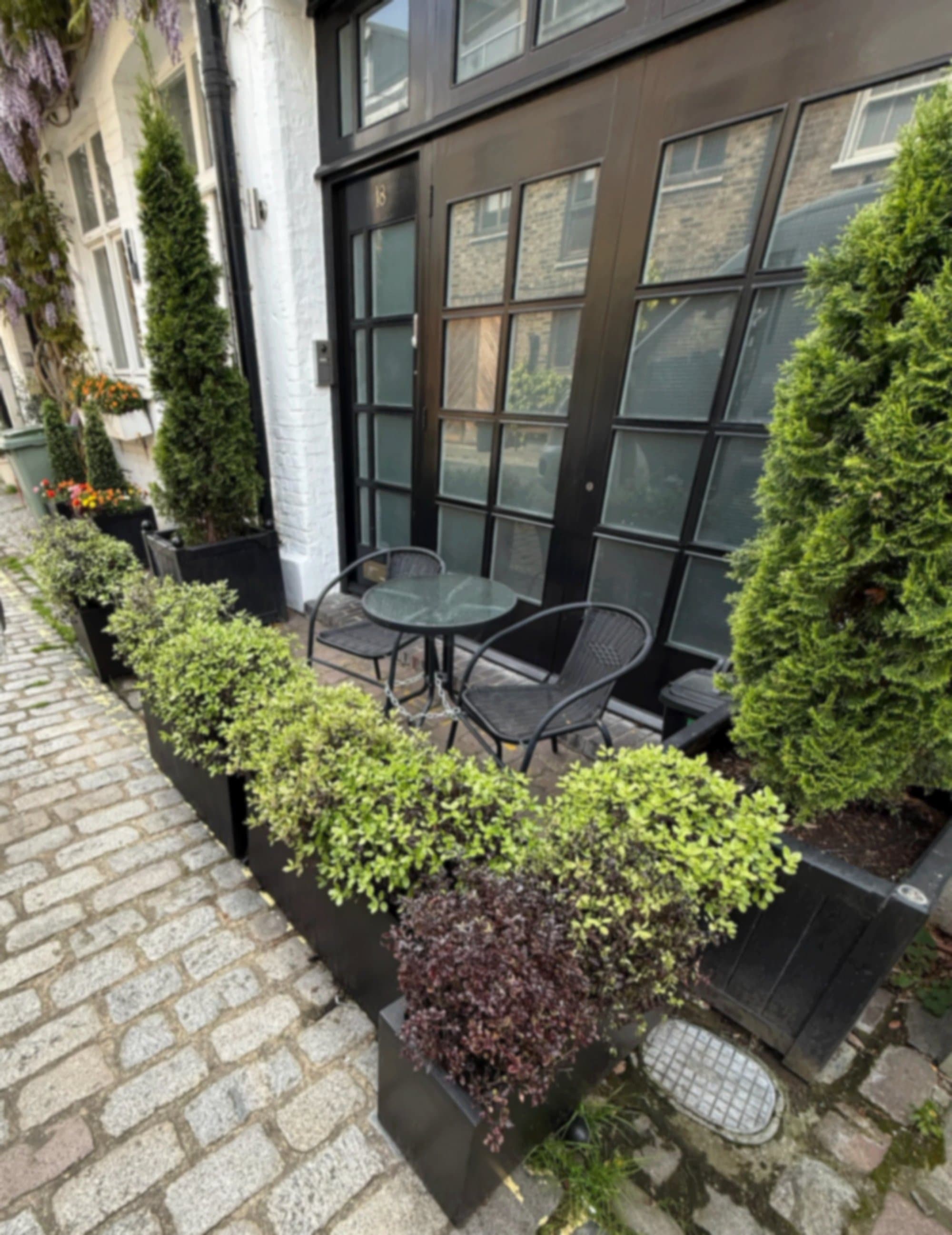The finished frontage of a Marylebone mews house with faux Buxus balls beneath the windows, tall dark planters framing a small seating area and a cobbled street underfoot