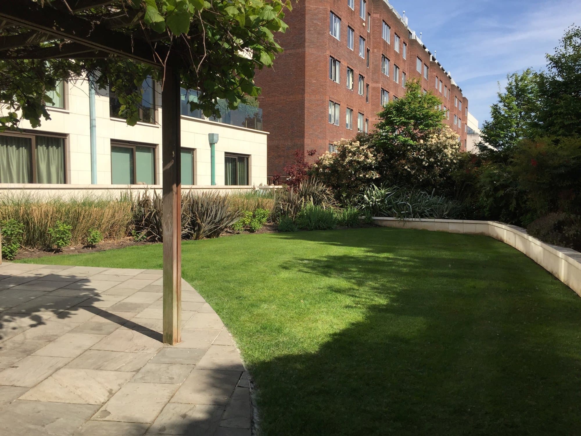 A neatly maintained communal garden beside a North London apartment block, a mown lawn edged with a pale stone retaining wall and planted borders, with a paved terrace under a pergola.