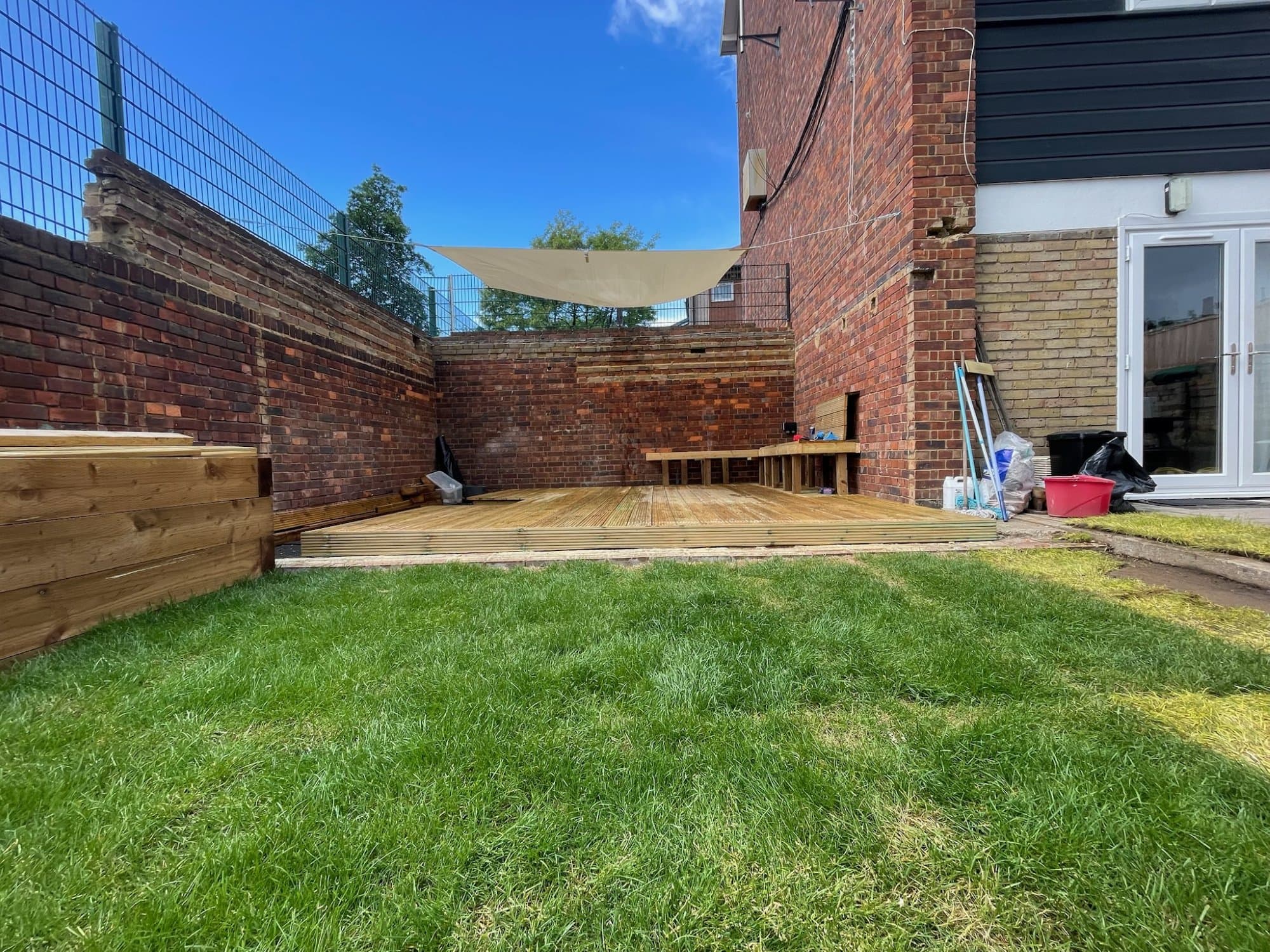 A newly built hardwood deck with a built-in bench and raised planter against exposed brick garden walls, a shade sail overhead and fresh lawn in front, on a North London project.